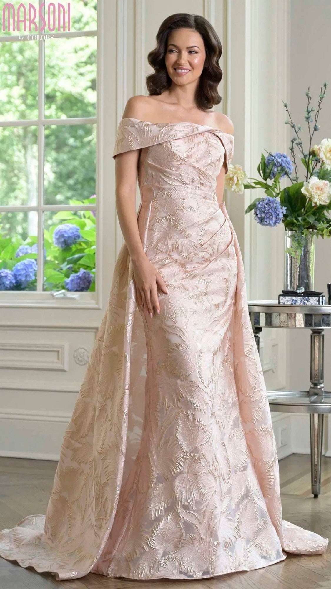 A woman stands indoors in the Marsoni MV1382 long formal dress, featuring an off-the-shoulder blush floral design. Large windows, white walls, and vases of flowers create an elegant backdrop for this sophisticated evening look.